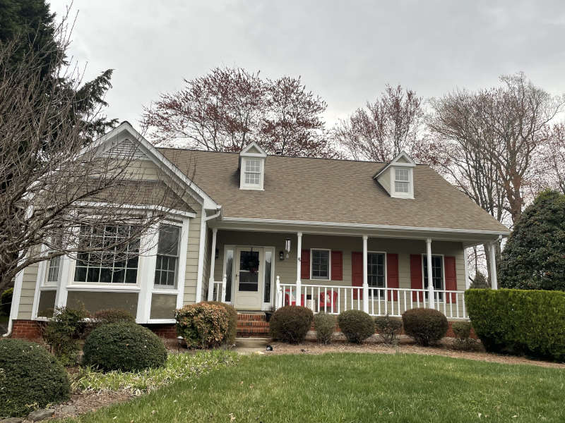 A cozy beige house with a front porch sits quietly among trees beneath a cloudy sky.