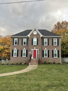 A charming brick house stands amid autumn trees, its red door and porch welcoming on a lush green lawn.